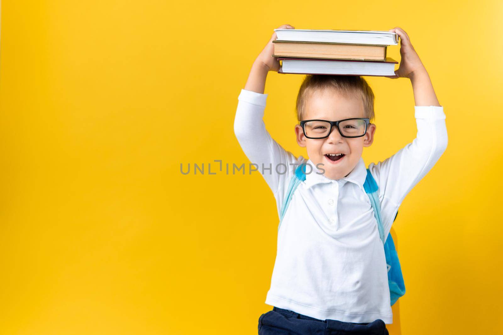 Banner Funny Preschool Child Boy in Glasses with Book on Head and Bag on Yellow Background Copy Space. Happy smiling kid go back to school, kindergarten. Success, motivation, winner, genius concept