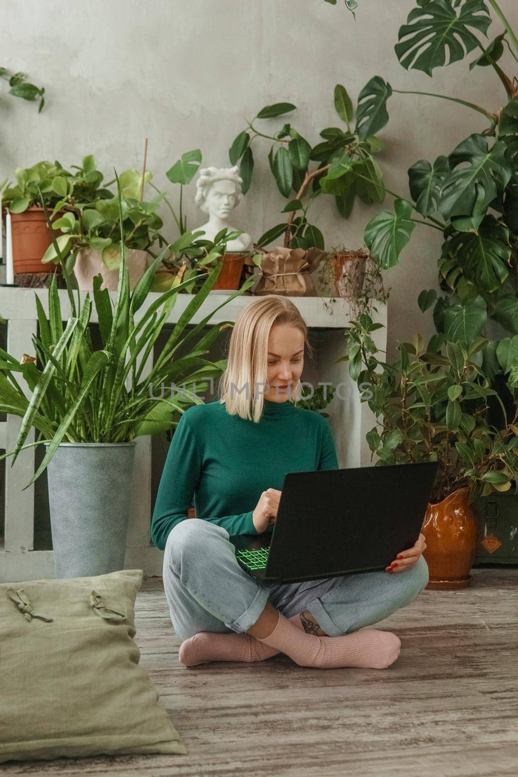 A blonde woman in a room with a lot of green indoor plants is working on a laptop. The concept of biophysical design in the interior. Work from home, work as a freelancer.