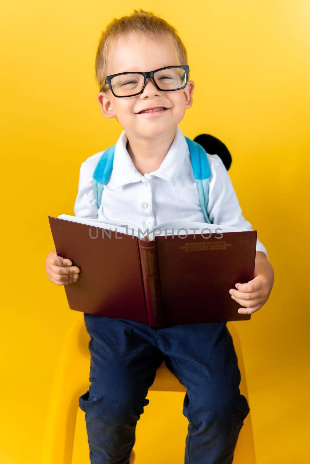Portrait of Funny Preschool Child Boy in Big Glasses sits on chair Reads Book on Yellow Background. Happy Smiling Kid Go Back to School, Kindergarten. Success, Motivation, Genius, Superhero concept.
