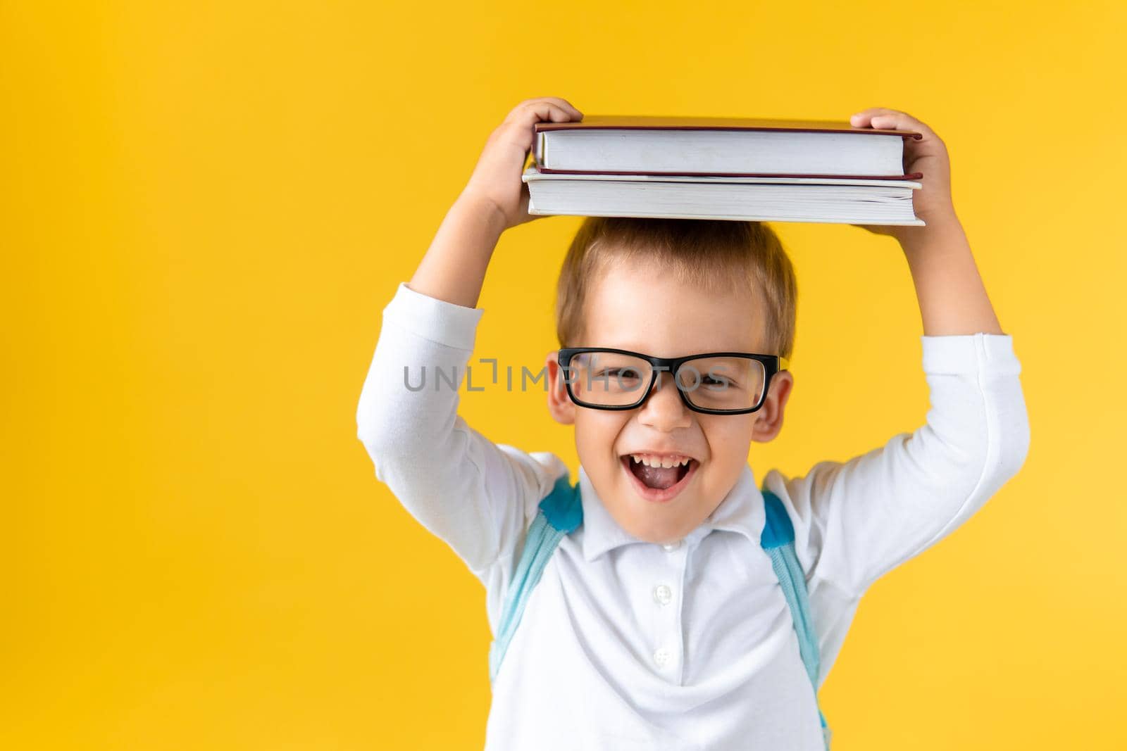 Funny Preschool Child Boy in Glasses with Book on Head and Bag on Yellow Background Copy Space. Happy Smiling Kid Go Back to School, Kindergarten. Success, Motivation, Winner Genius, Superhero concept.