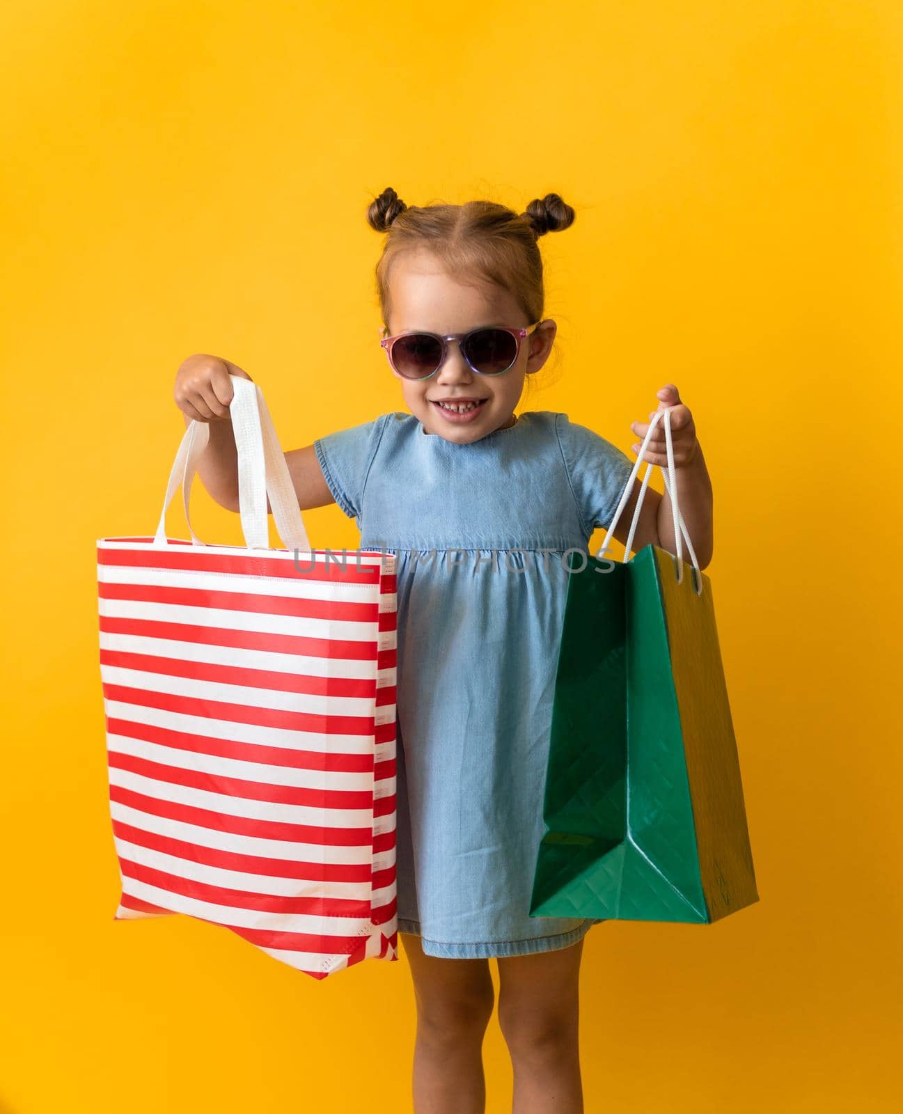 Portrait Beautiful Happy Little Preschool Girl In Sunglasses Smiling Cheerful Holding Cardboard Bags Isolated On Orange Yellow Studio background. Happiness, Consumerism, Sale People shopping Concept.