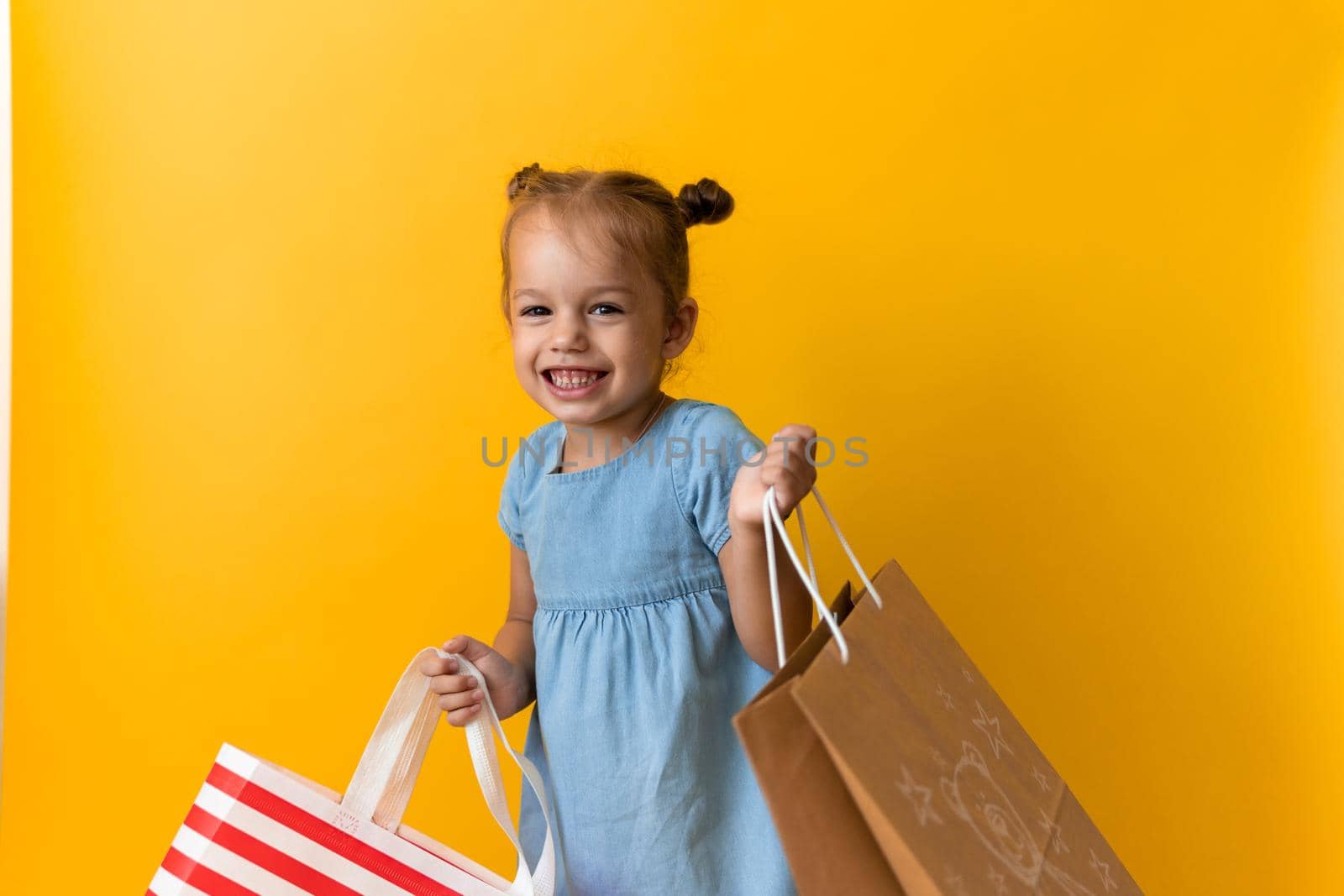 Portrait Caucasian Beautiful Happy Little Preschool Girl Smiling Cheerful And Holding Cardboard Bags Isolated On Orange Yellow Studio background. Happiness, Consumerism, Sale People shopping Concept