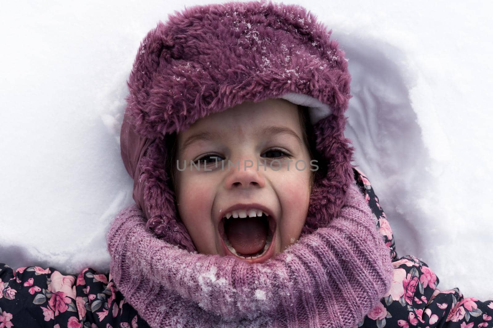 Winter, family, childhood concepts - close-up portrait authentic little preschool girl in pink clothes smile laugh shout with open mouth laying on snow in frosty weather day outdoors. Funny kid face.