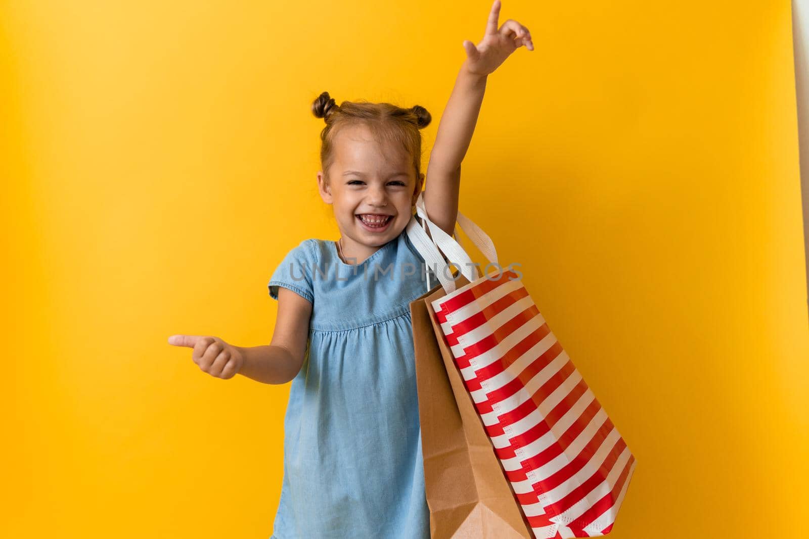 Portrait Beautiful Happy Little Preschool Girl Smiling Cheerful Holding Cardboard Bags Points Finger To Side And Up On Orange Yellow Background. Happiness, Consumerism, Sale People Shopping Concept.