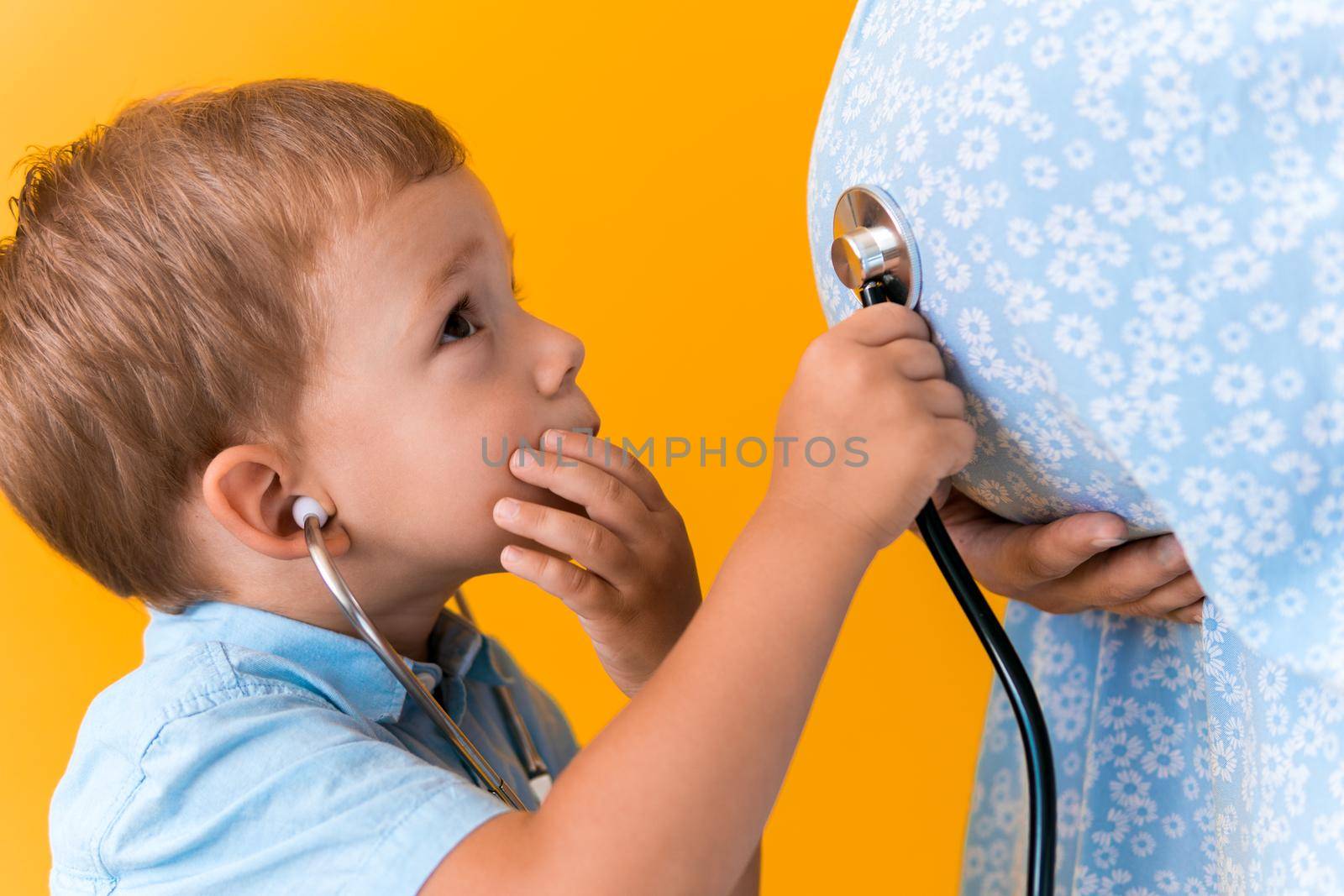 Motherhood, hot summer - croped portrait pregnant mother unrecognizable woman blue dress little boy son sibling treat pregnant mom role play stethoscope unborn brother mom tummy on yellow background.