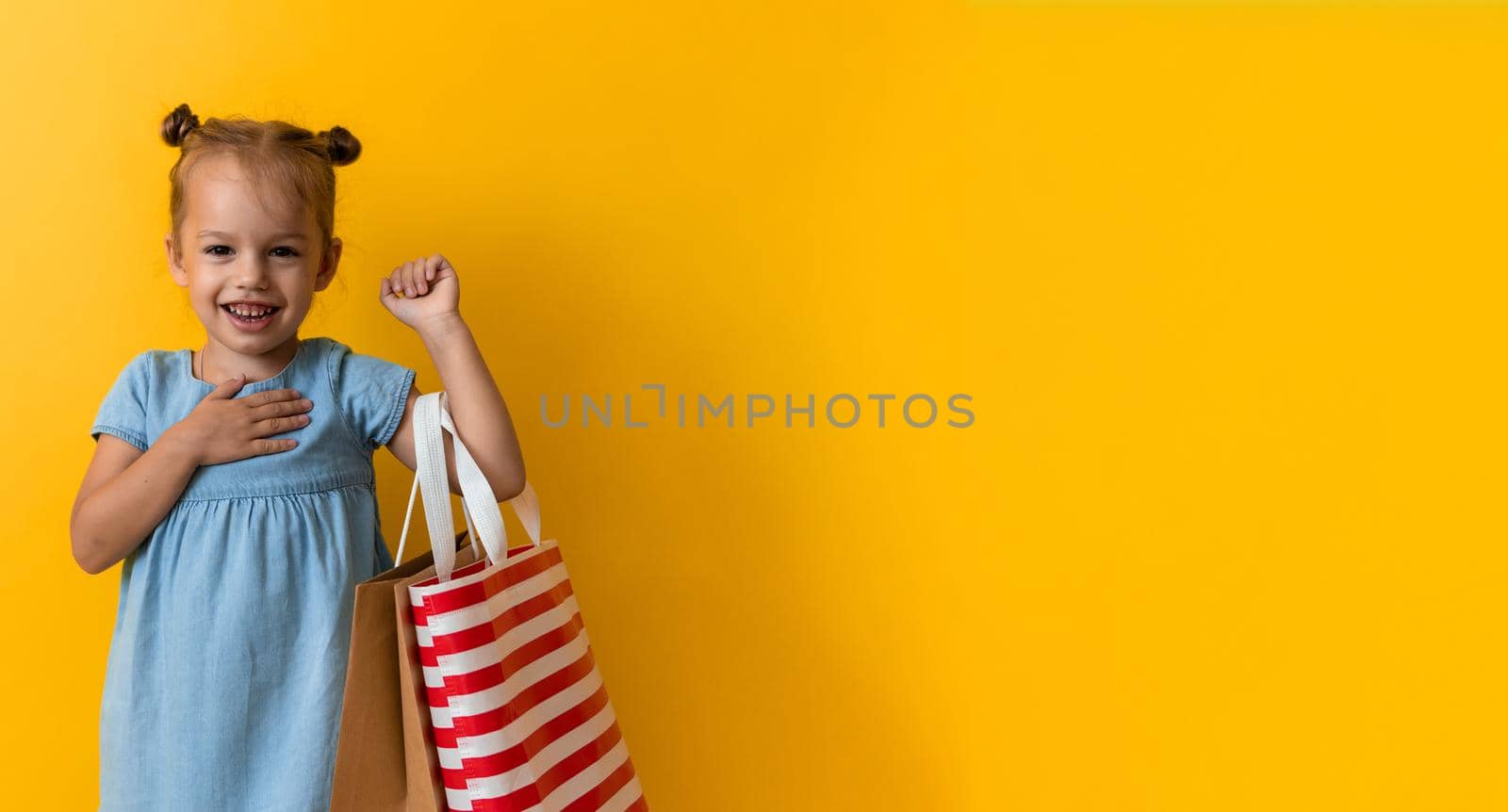 Banner Portrait Caucasian Beautiful Happy Little Preschool Girl Smiling Cheerful And Holding Cardboard Bags Isolated On Orange Yellow Background. Happiness, Consumerism, Sale People shopping Concept.