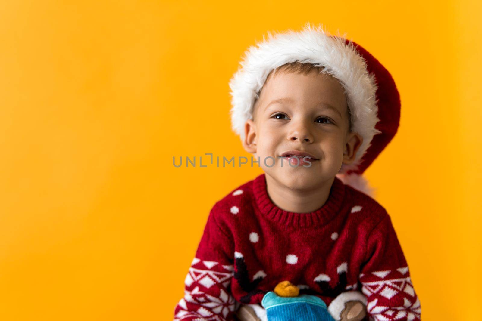 Portrait of happy smiling positive joyful positive preschool little boy in red warm santa hat showing thumb up on orange, yellow background. Winter, holiday, celebration, Christmas, New Year copy space.