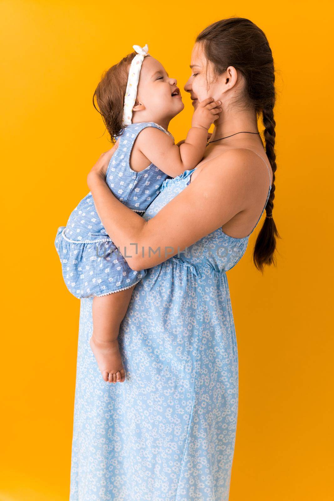 Motherhood, hot summer - portrait young pregnant happy smiling joy mother woman in blue dress holding little baby daughter toddler sibling bare feet in arms looking into eyes kiss on yellow background by mytrykau