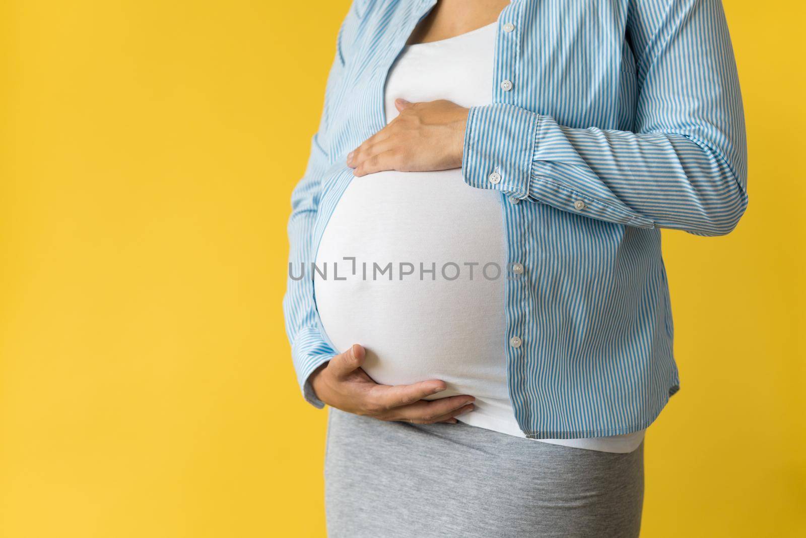 Motherhood, femininity, love, care, waiting, hot summer - bright croped Close-up unrecognizable pregnant woman in shirt with small baby shoes hand over tummy rub belly on yellow background, copy space.