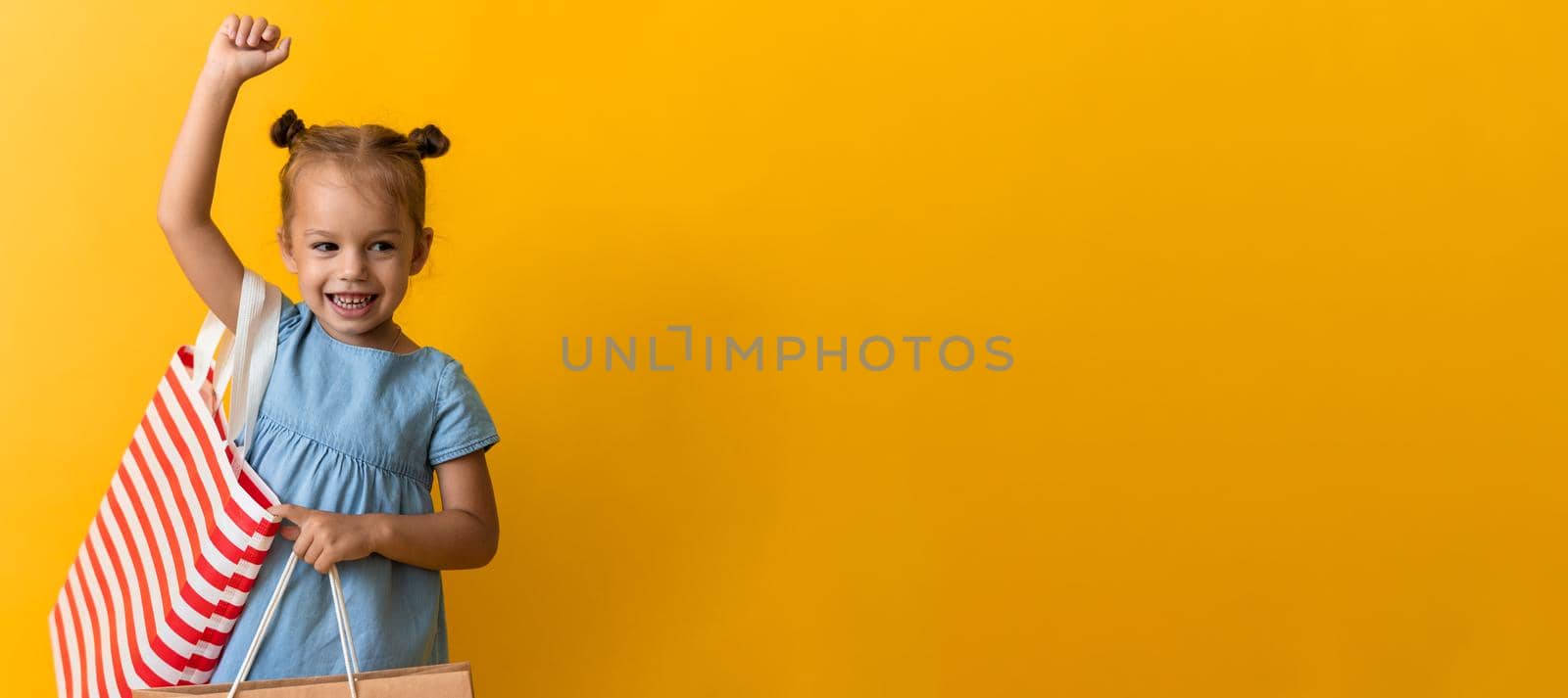 Banner Portrait Caucasian Beautiful Happy Little Preschool Girl Smiling Cheerful And Holding Cardboard Bags Isolated On Orange Yellow Background. Happiness, Consumerism, Sale People shopping Concept.