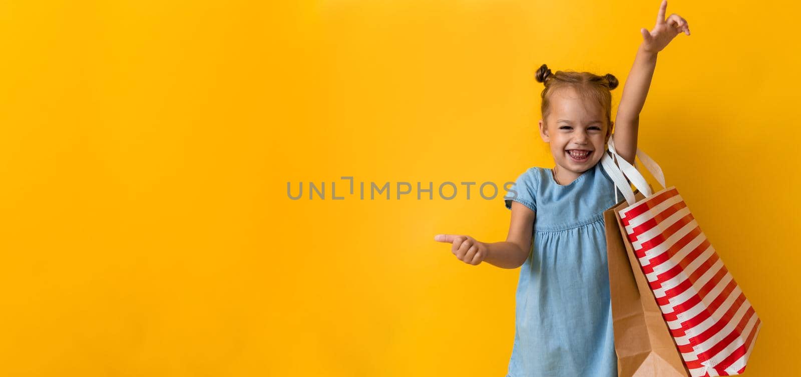 Banner Portrait Beautiful Happy Little Preschool Girl Smiling Cheerful Holding Cardboard Bags Points Finger To Side Up On Orange Yellow Background. Happiness, Consumerism, Sale People shopping Concept by mytrykau