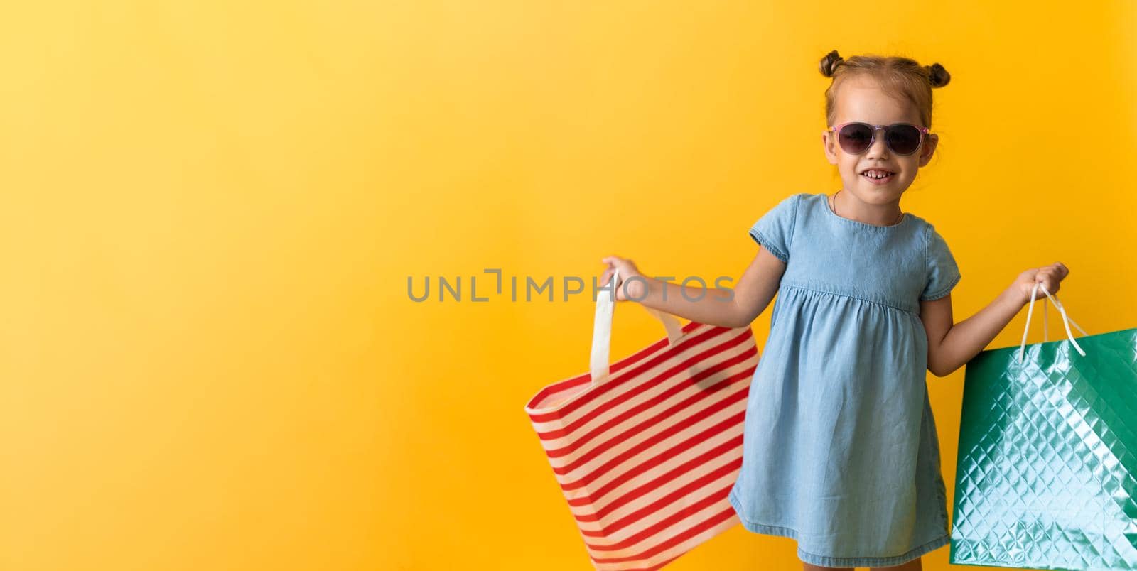 Portrait Beautiful Happy Little Preschool Girl In Sunglasses Smiling Cheerful Holding Cardboard Bags Isolated On Orange Yellow Studio background. Happiness, Consumerism, Sale People shopping Concept.