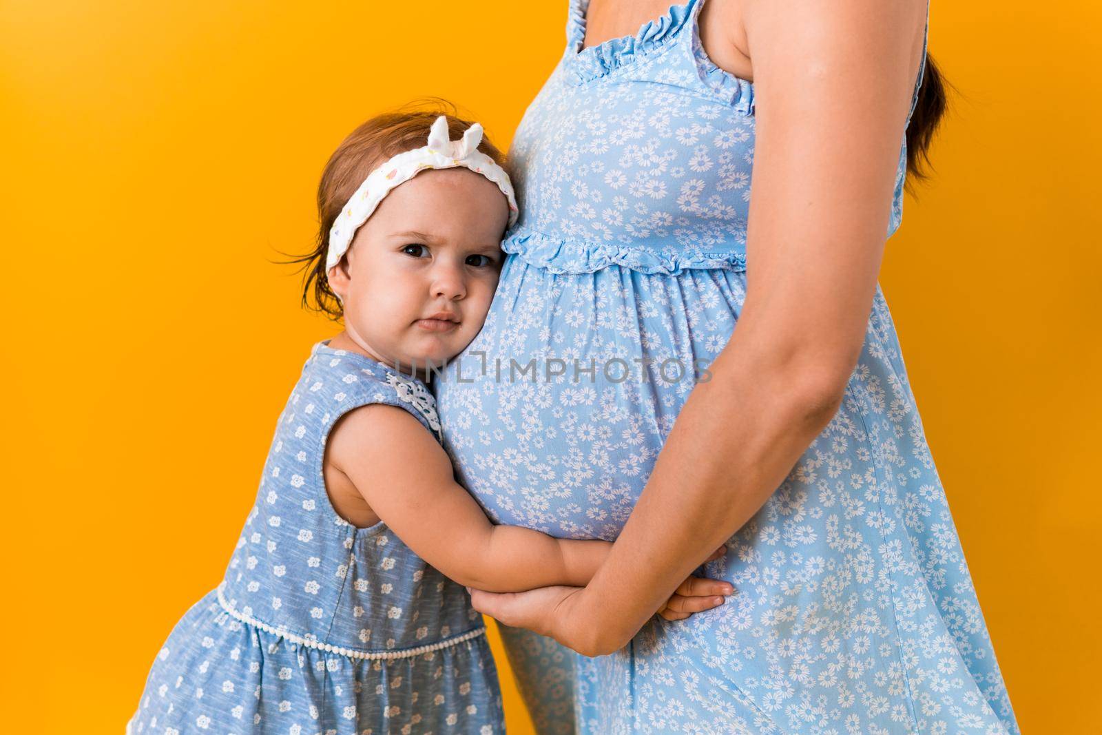 Motherhood, love, Childhood, hot summer - croped portrait pregnant mother unrecognizable woman blue dress little sibling girl sister hug touch mum big belly brother on yellow background copy space.