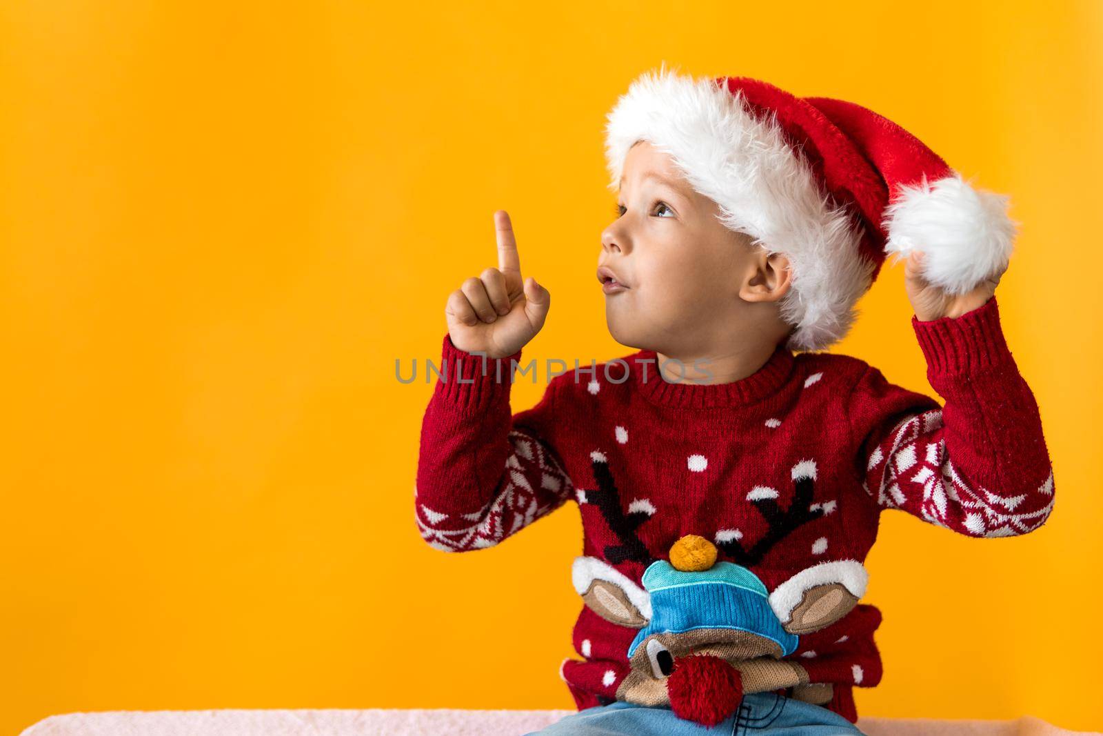 Portrait of happy smiling positive joyful positive preschool little boy in red warm santa hat showing thumb up on orange, yellow background. Winter, holiday, celebration, Christmas, New Year copy space by mytrykau