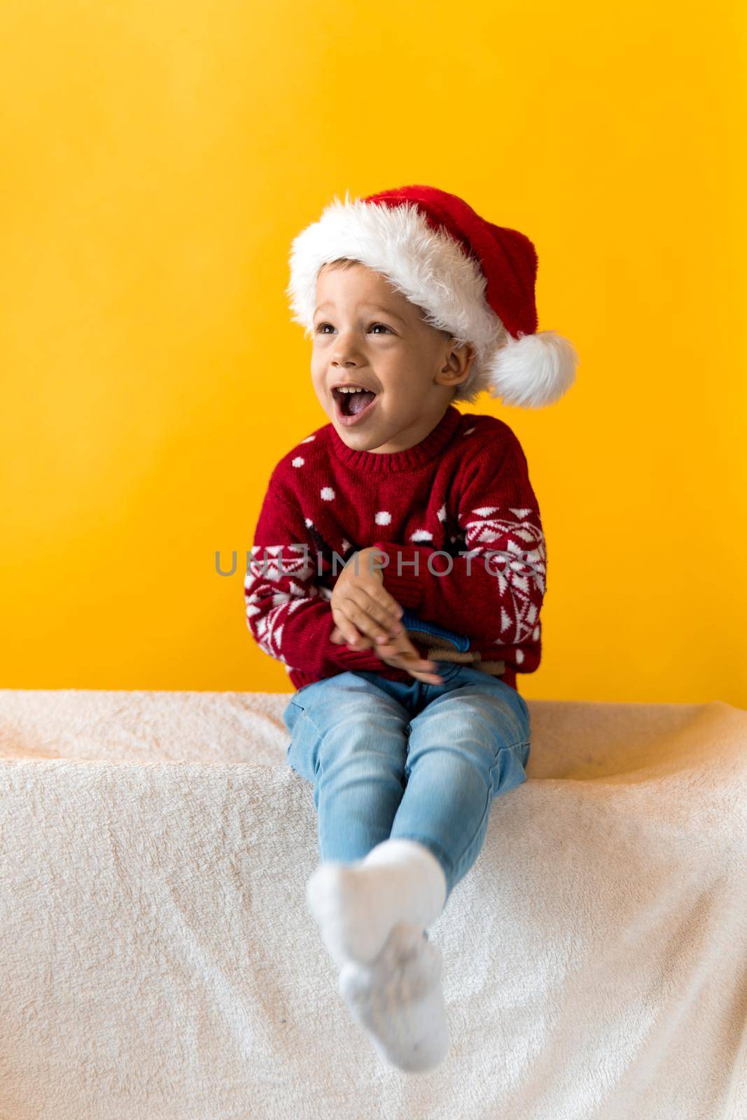 Portrait of happy smiling positive joyful positive preschool little boy in red warm santa hat showing thumb up on orange, yellow background. Winter, holiday, celebration, Christmas, New Year copy space by mytrykau