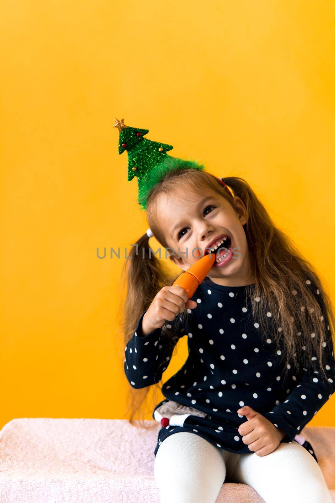 Portrait positive cheerful smiling happy little schoolgirl girl Christmas tree decoration polka dot dress biting eat orange carrots on orange background. New year, holiday, celebration, winter concept.