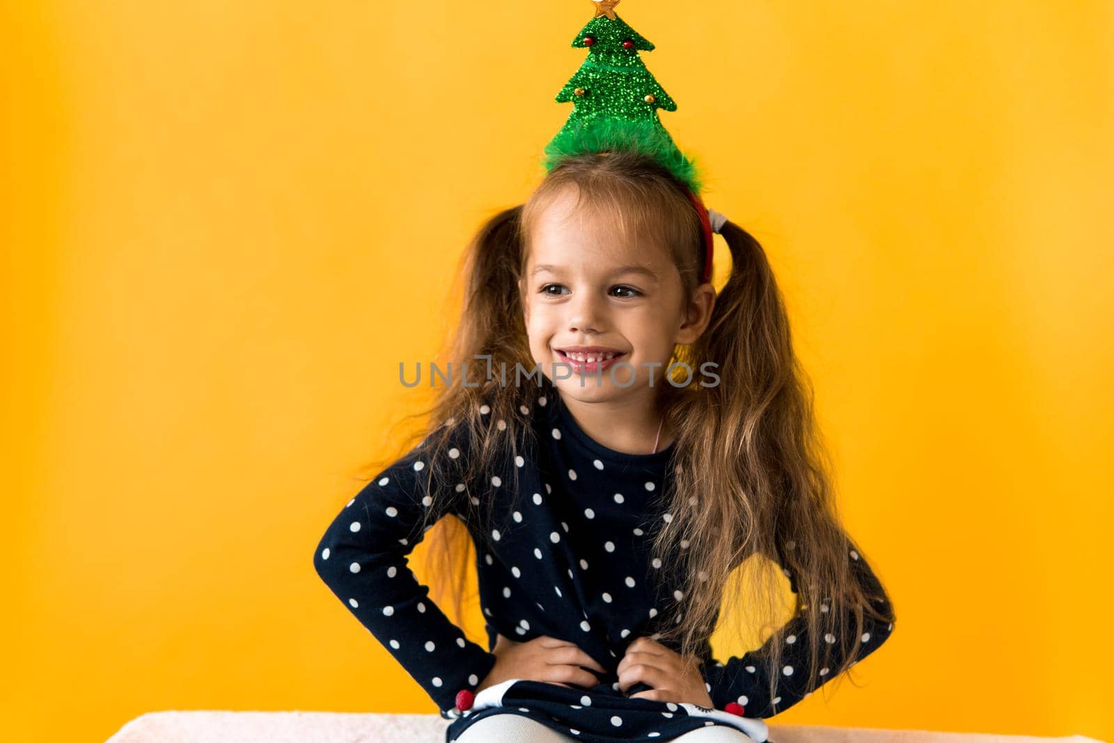 Portrait of positive smiling surprised happy little schoolgirl girl Christmas tree decoration on head polka dot dress show thumbs up orange background. New year, holiday, celebration, winter concept.