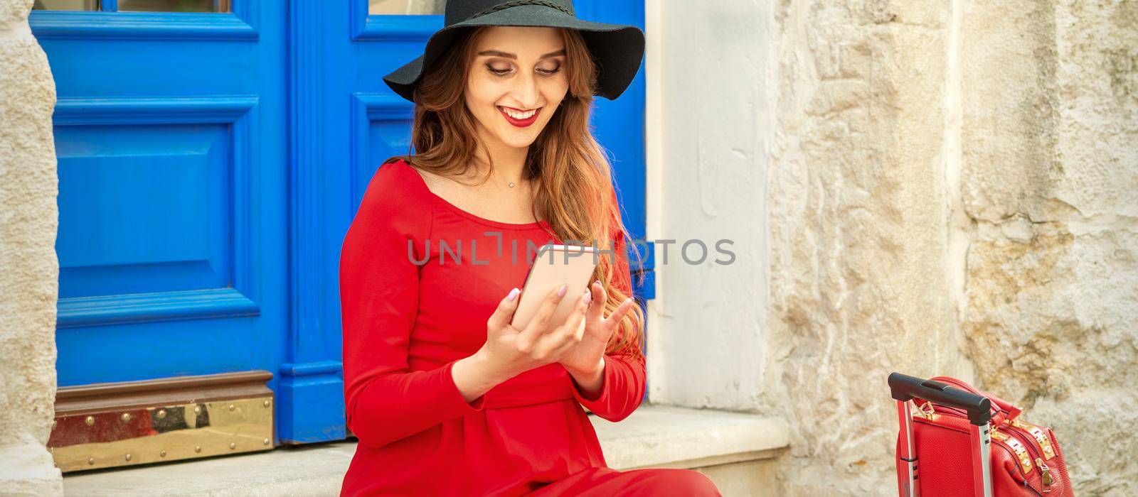 Beautiful young caucasian traveling woman in black hat looking on the smartphone smiling and sitting on stairs at the door outdoors