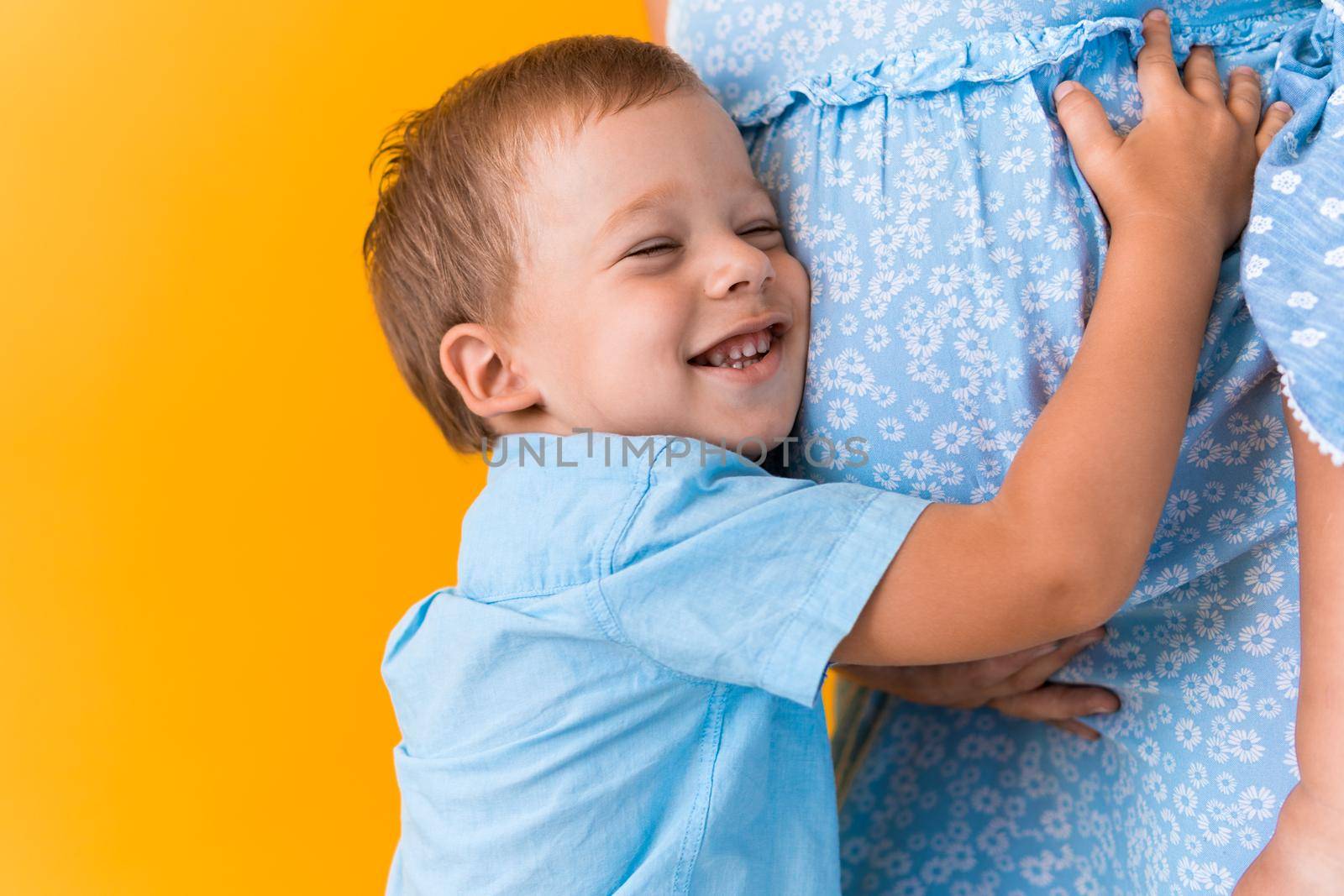 Motherhood, femininity, love, Childhood, hot summer - croped portrait pregnant unrecognizable woman blue dress little preschool boy son sibling hug mum big belly brother on yellow background copy space.