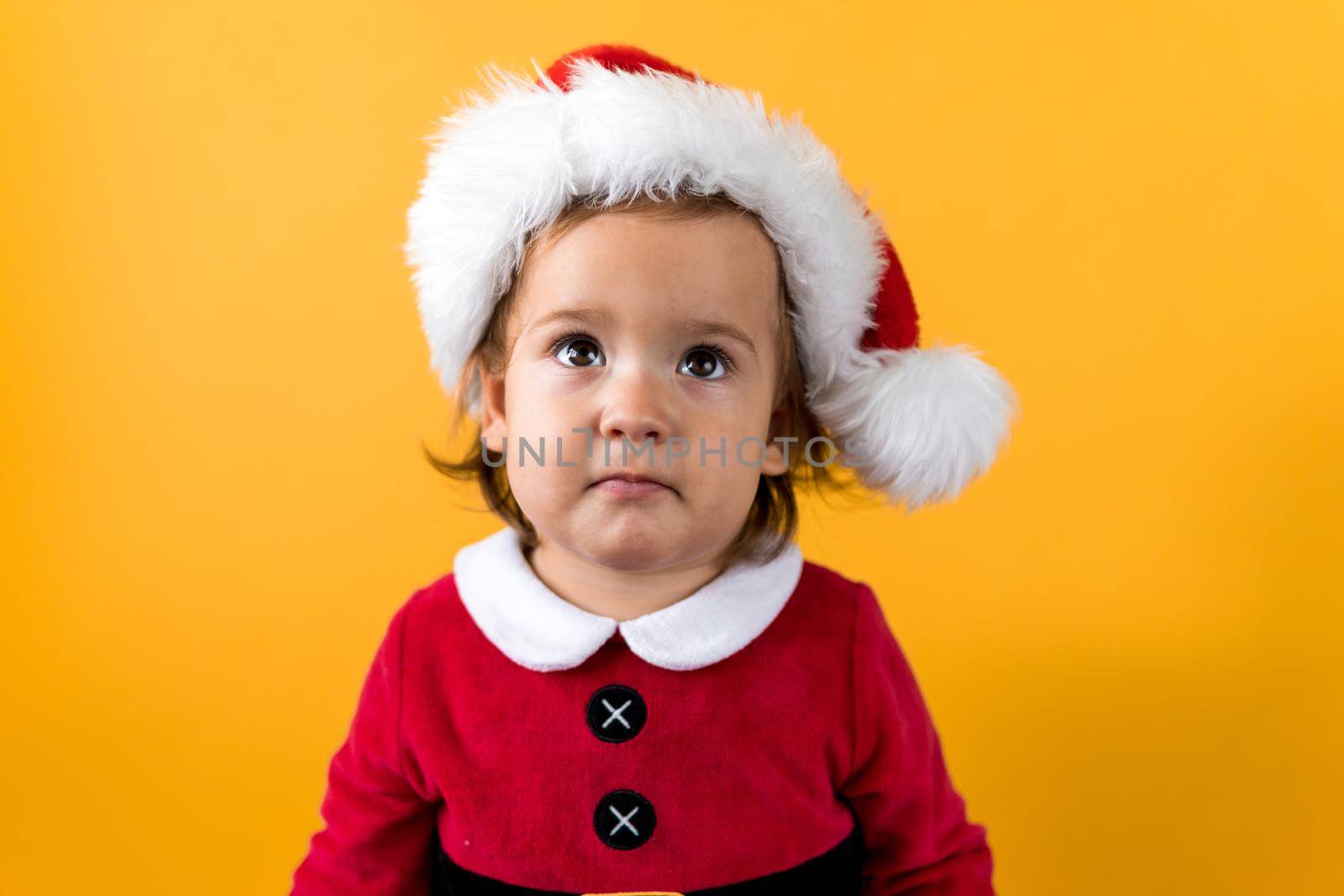 Smiling Portraite Cute Cheerful Chubby Baby Girl in Santa Hat Looking Up Interested At Yellow Background. Child Play Christmas Scene Celebrating Birthday. Kid Have Fun Spend New Year Time Copy Space.
