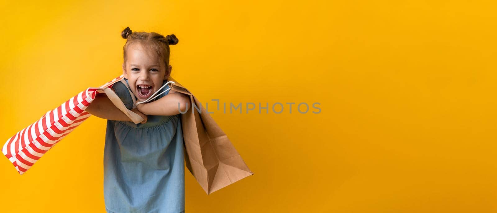 Banner Portrait Caucasian Beautiful Happy Little Preschool Girl Smiling Cheerful And Holding Cardboard Bags Isolated On Orange Yellow Background. Happiness, Consumerism, Sale People shopping Concept by mytrykau