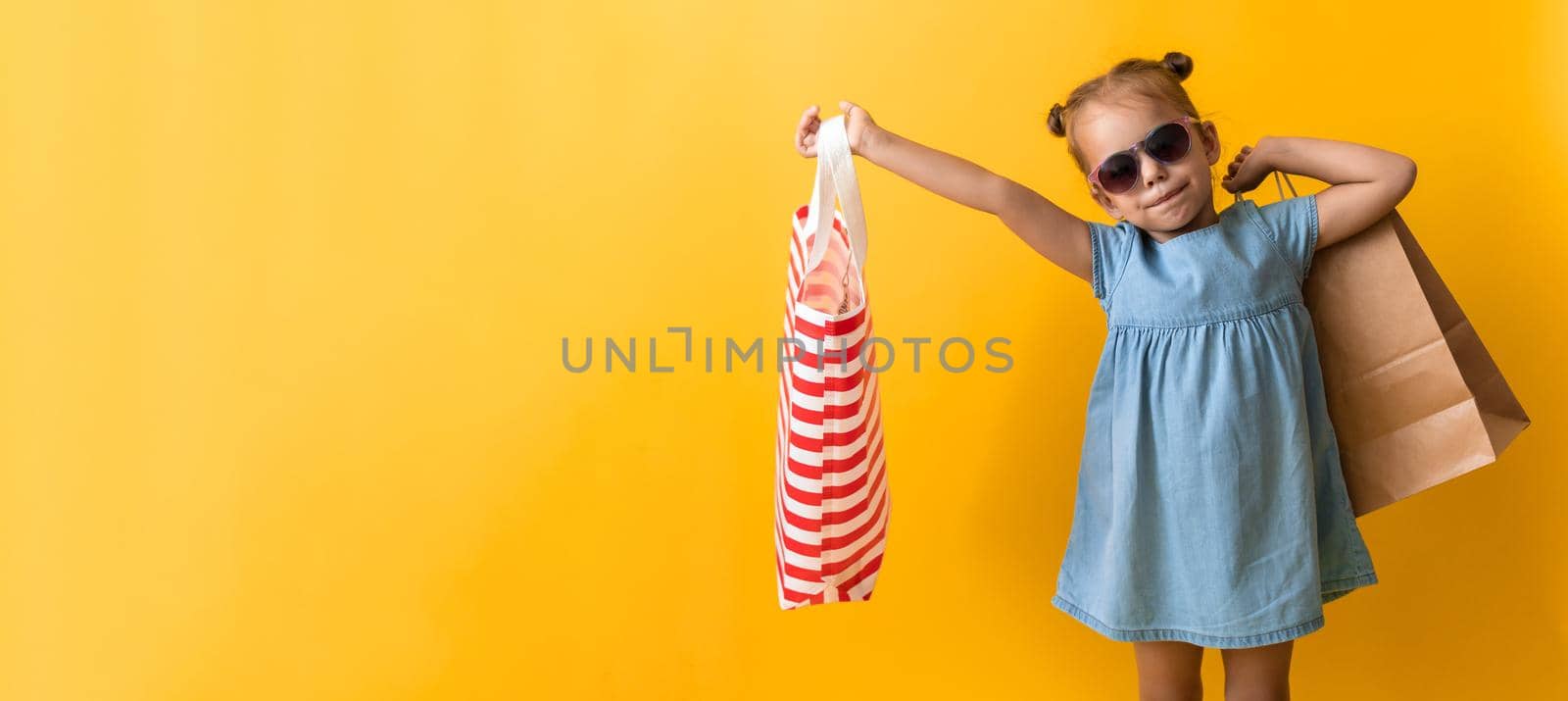 Banner Portrait Beautiful Happy Little Preschool Girl In Sunglasses Smiling Cheerful Holding Cardboard Bags Isolated On Orange Yellow Background. Happiness, Consumerism, Sale People shopping Concept by mytrykau