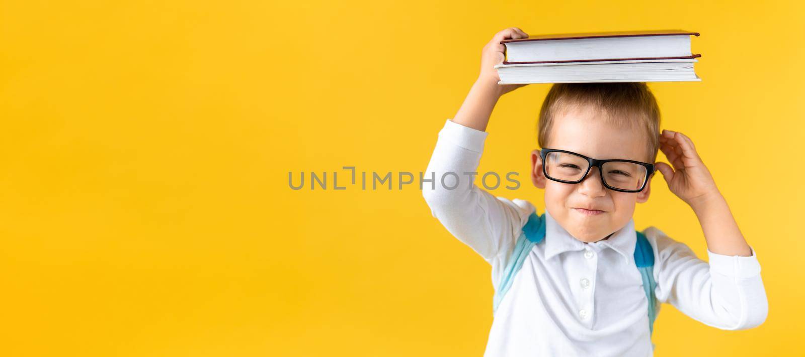 Banner Funny Preschool Child Boy in Glasses with Book on Head and Bag on Yellow Background Copy Space. Happy smiling kid go back to school, kindergarten. Success, motivation, winner, genius concept