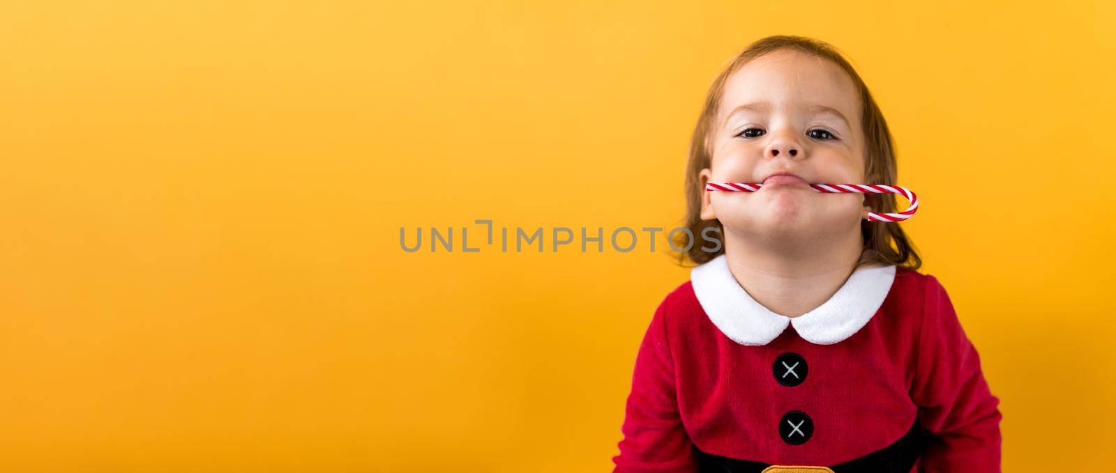 Banner Portraite Cute Cheerful Chubby Baby Girl in Santa Suite Holding Eating Caramel Candy At Yellow Background. Child Play Christmas Celebrating Birthday. Kid Have Fun Spend New Year Time Copy Space.