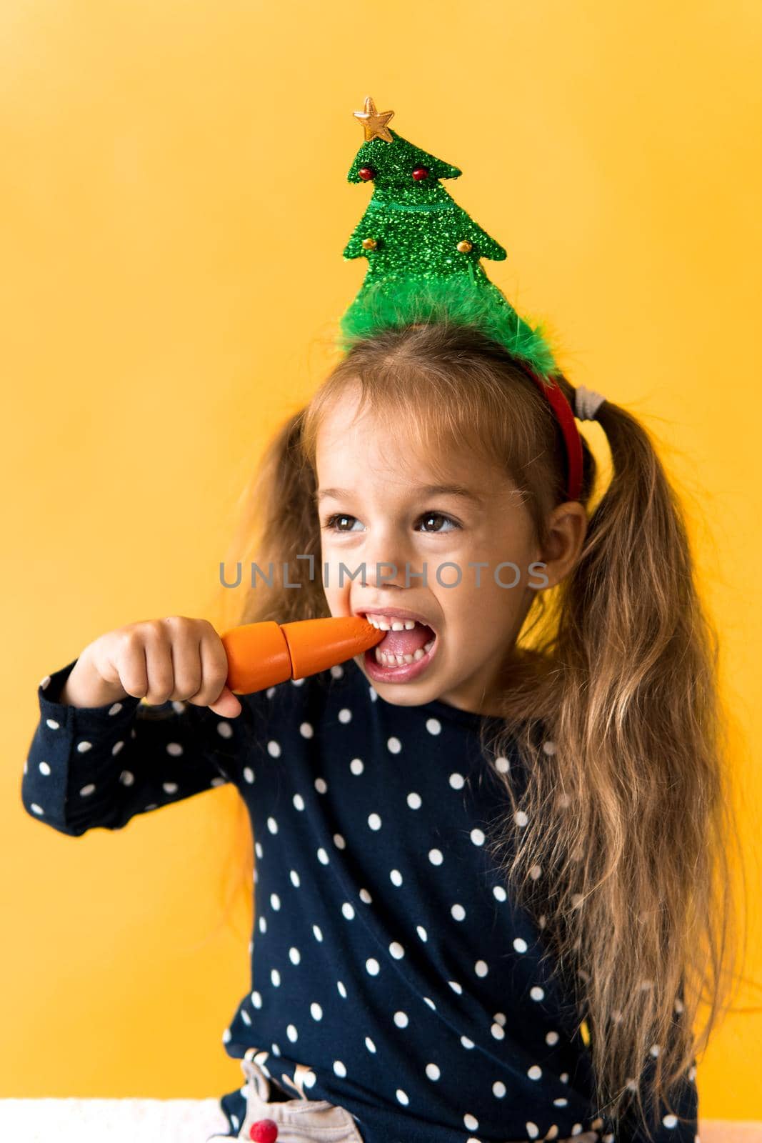 Portrait positive cheerful smiling happy little schoolgirl girl Christmas tree decoration polka dot dress biting eat orange carrots on orange background. New year, holiday, celebration, winter concept.