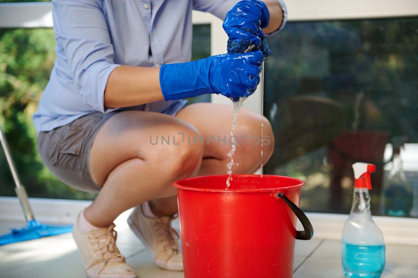 Details: Hands in blue rubber work gloves of a maid squeezing a cloth into a red bucket during a general cleaning of a house and veranda. Housekeeping. Housework. Chores. Professional cleaning service