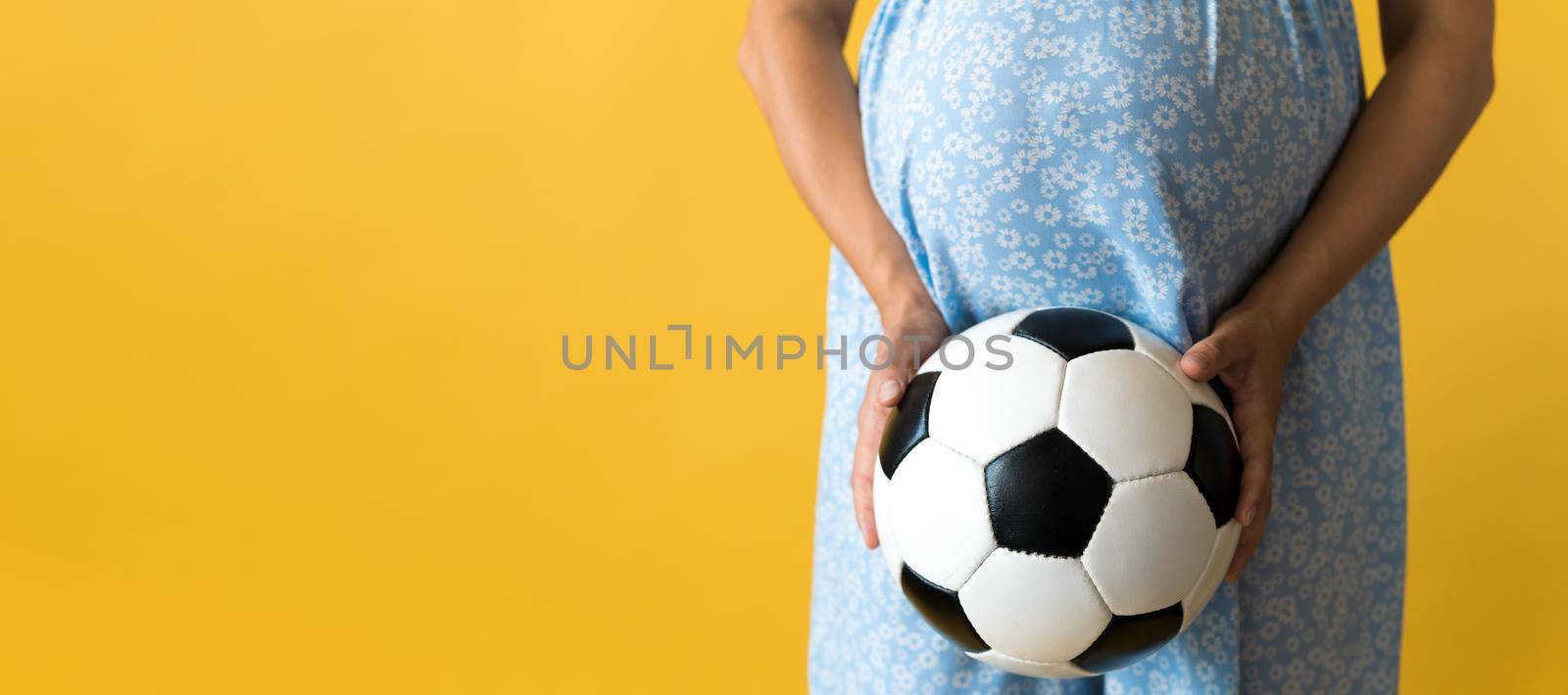 Motherhood, femininity, football, sport, dairy, hot summer. banner pregnant young pretty woman in floral blue dress holds soccer white and black leather classic ball rubs tummy on yellow background by mytrykau