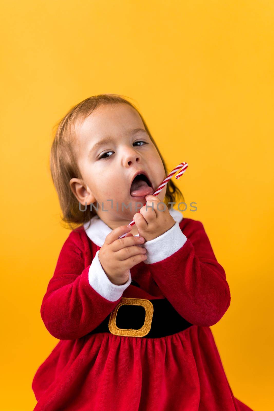 Portraite Cute Cheerful Chubby Baby Girl in Santa Suite Holding Eating Caramel Candy At Yellow Background. Child Play Christmas Scene Celebrating Birthday. Kid Have Fun Spend New Year Time Copy Space.