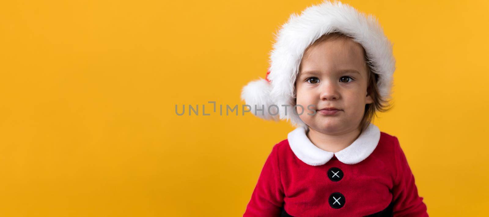 Banner Portraite Cute Happy Cheerful Chubby Baby Girl in Santa Suit Looking On Camera At Yellow Background. Child Play Christmas Scene Celebrating Birthday. Kid Have Fun Spend New Year Time Copy Space by mytrykau