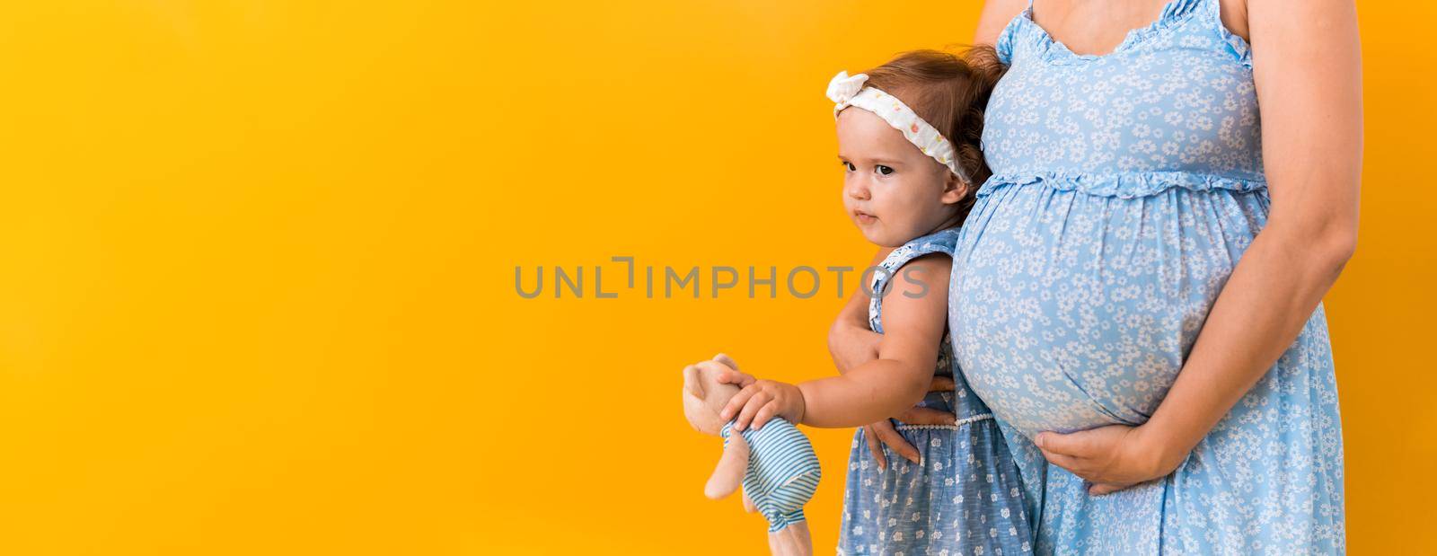 Motherhood, femininity, hot summer - croped banner pregnant unrecognizable woman mother in blue dress little happy girl baby sibling with teddy bear hugs care mom's big belly on yellow background by mytrykau
