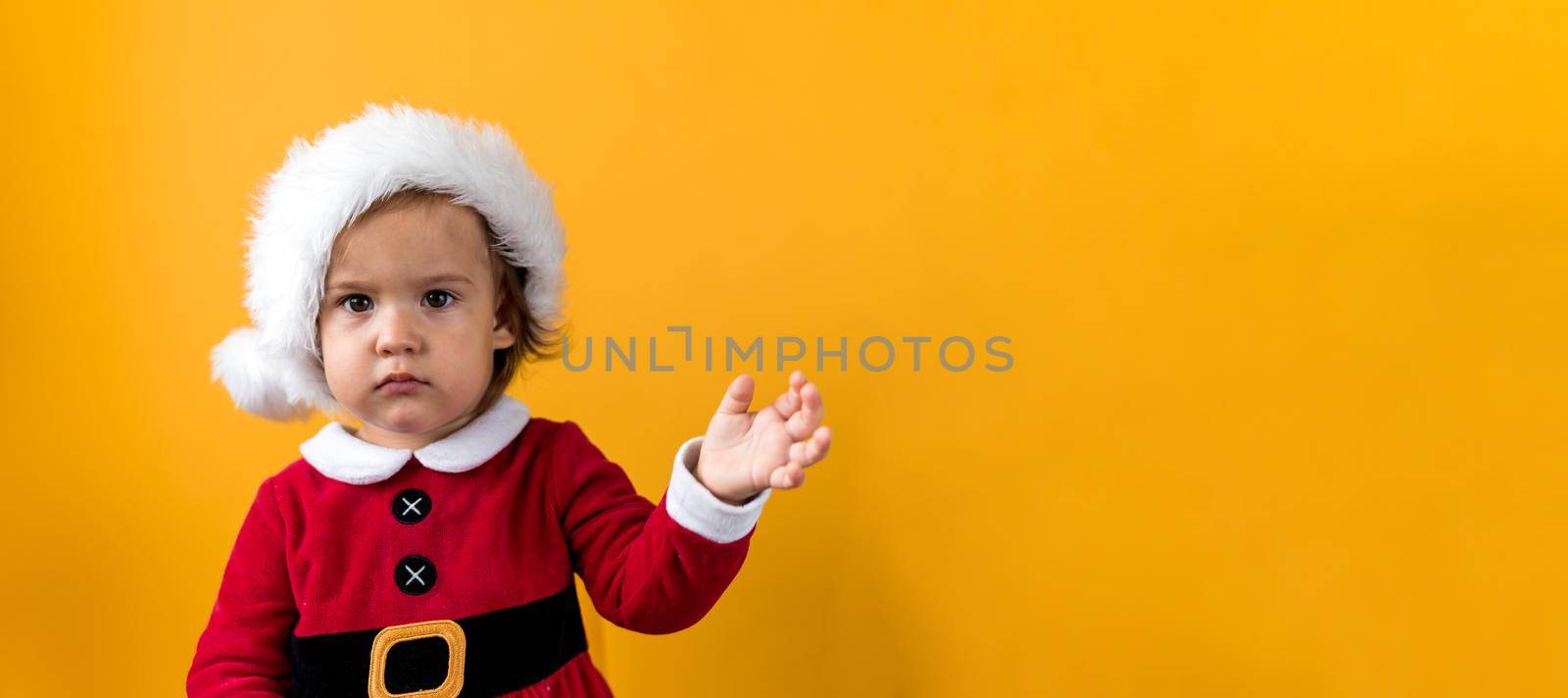 Banner Serious Portraite Cute Fun Cheerful Chubby Baby Girl in Santa Hat Waving Hand At Yellow Background. Child Play Christmas Scene Celebrating Birthday. Kid Have Fun Spend New Year Time Copy Space by mytrykau