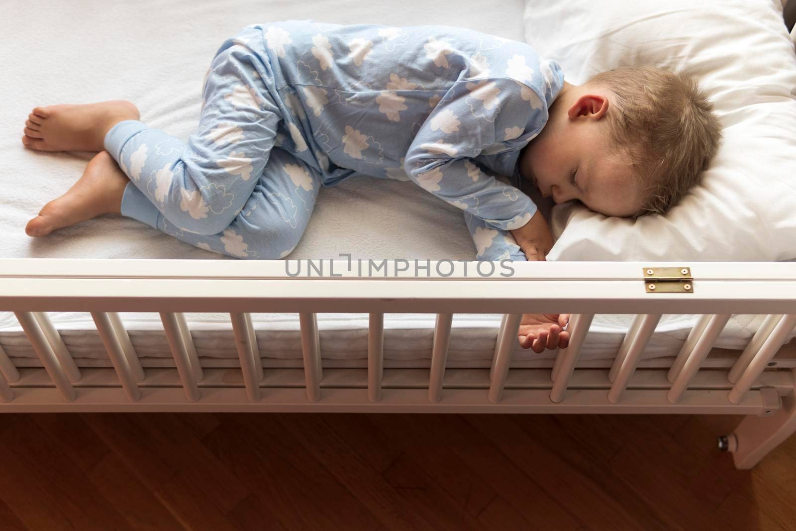top view Cute little 2-3 years preschool baby boyl kid sleeping sweetly in white crib during lunch rest time in blue pajama with pillow at home. Childhood, leisure, comfort, medicine, health concept.