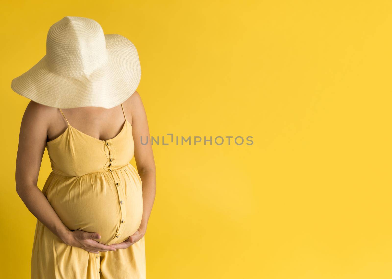 Motherhood, femininity, fashion, hot summer - banner unrecognizable pregnant woman lady in beautiful vintage dress Provence straw hat with hands over tummy rubs tummy on yellow background copy space.