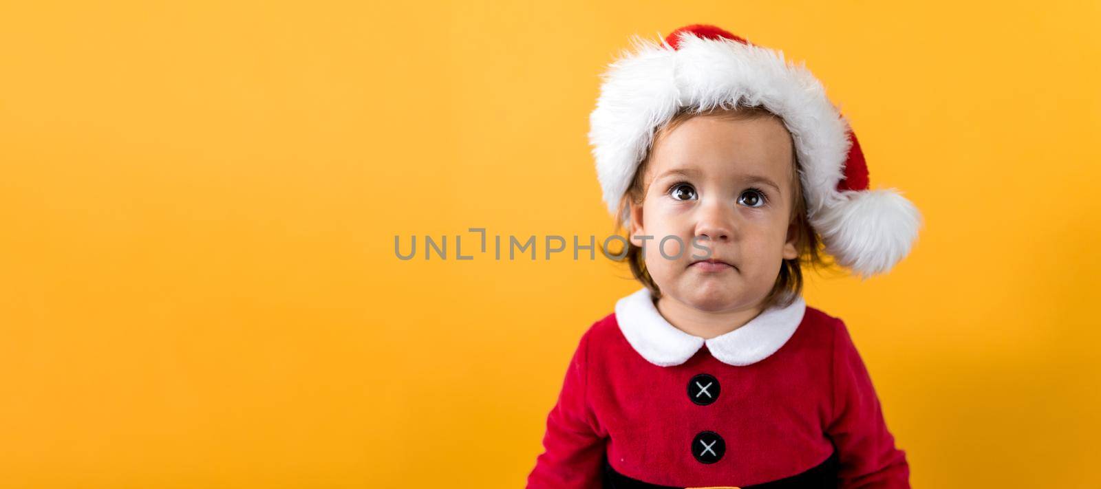 Cute Happy Cheerful Chubby Toddler Baby Girl in Santa Suit Looking On Camera At Yellow Background. Child Playing Scene Celebrating Birthday. Kid bites Christmas toy cookies decoration New Year Time.