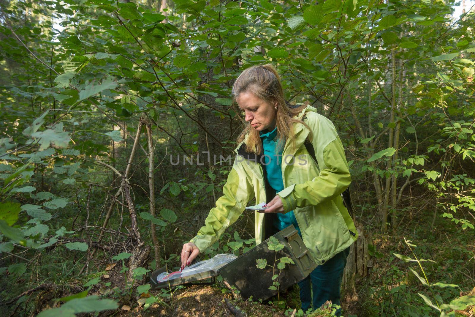 A woman in woods finds a large geocaching container.