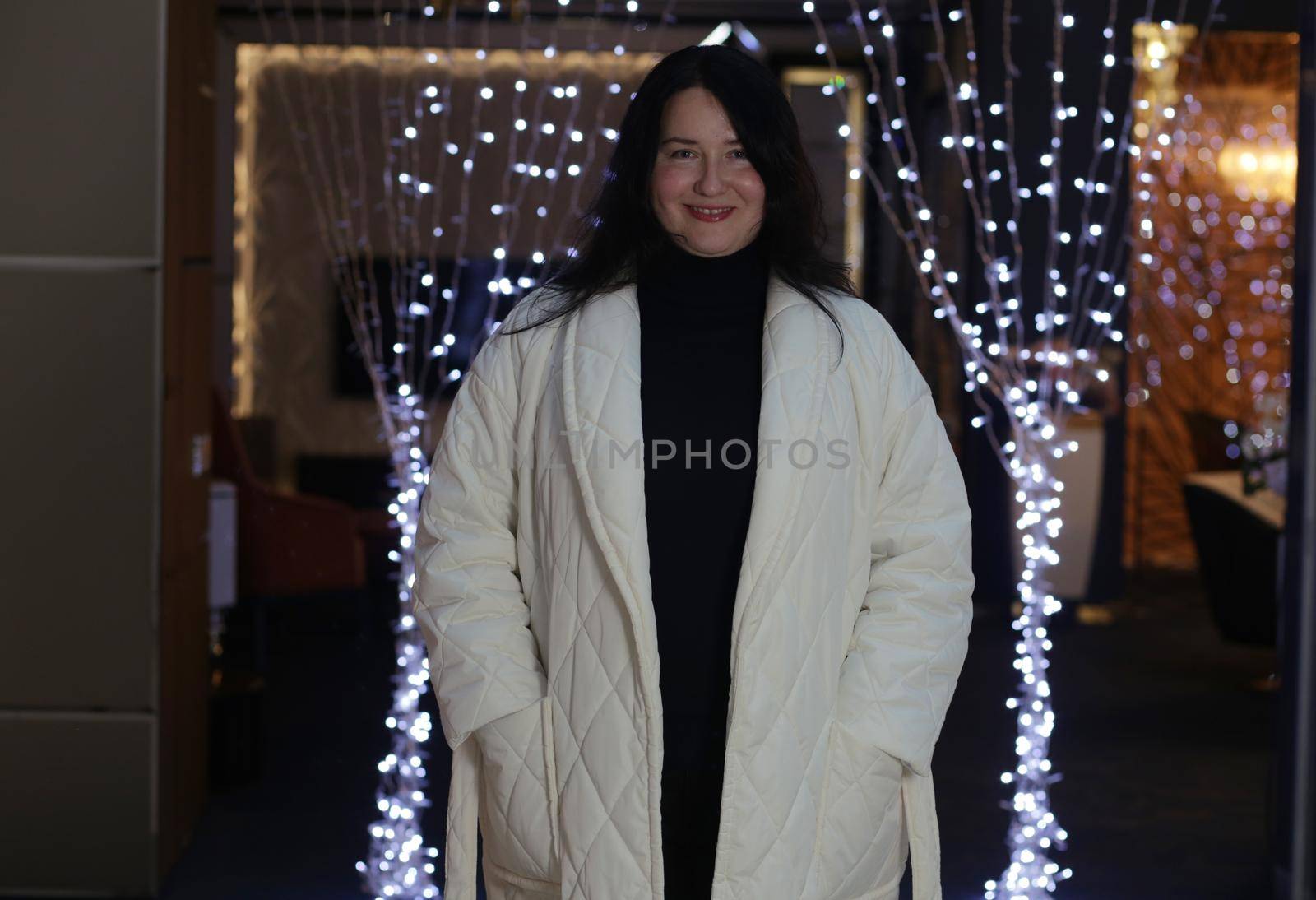 Cheerful brunette plus-size indoors in black sweater and white winter coat has her hands in her pockets and looks at the camera.