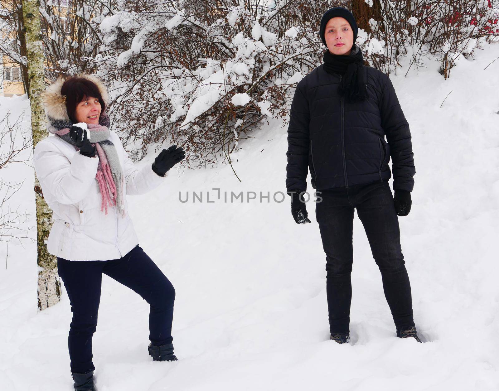 An elderly mother in a white jacket, in a scarf, holds a snowball in her hands near her son in black winter clothes against the backdrop of snow.