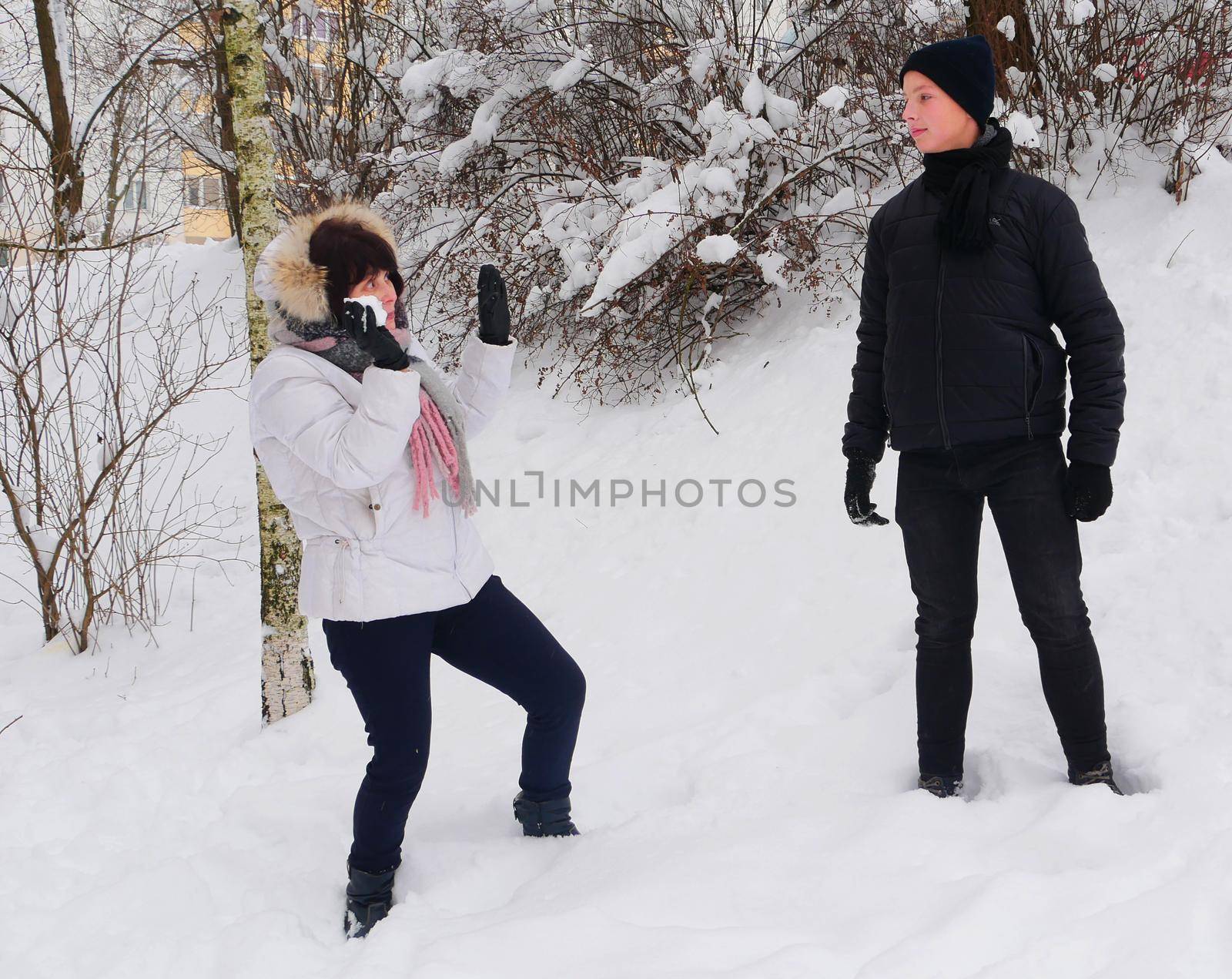 Mom in a white jacket and scarf throws a snowball at her joyful son against the backdrop of snow-covered bushes.