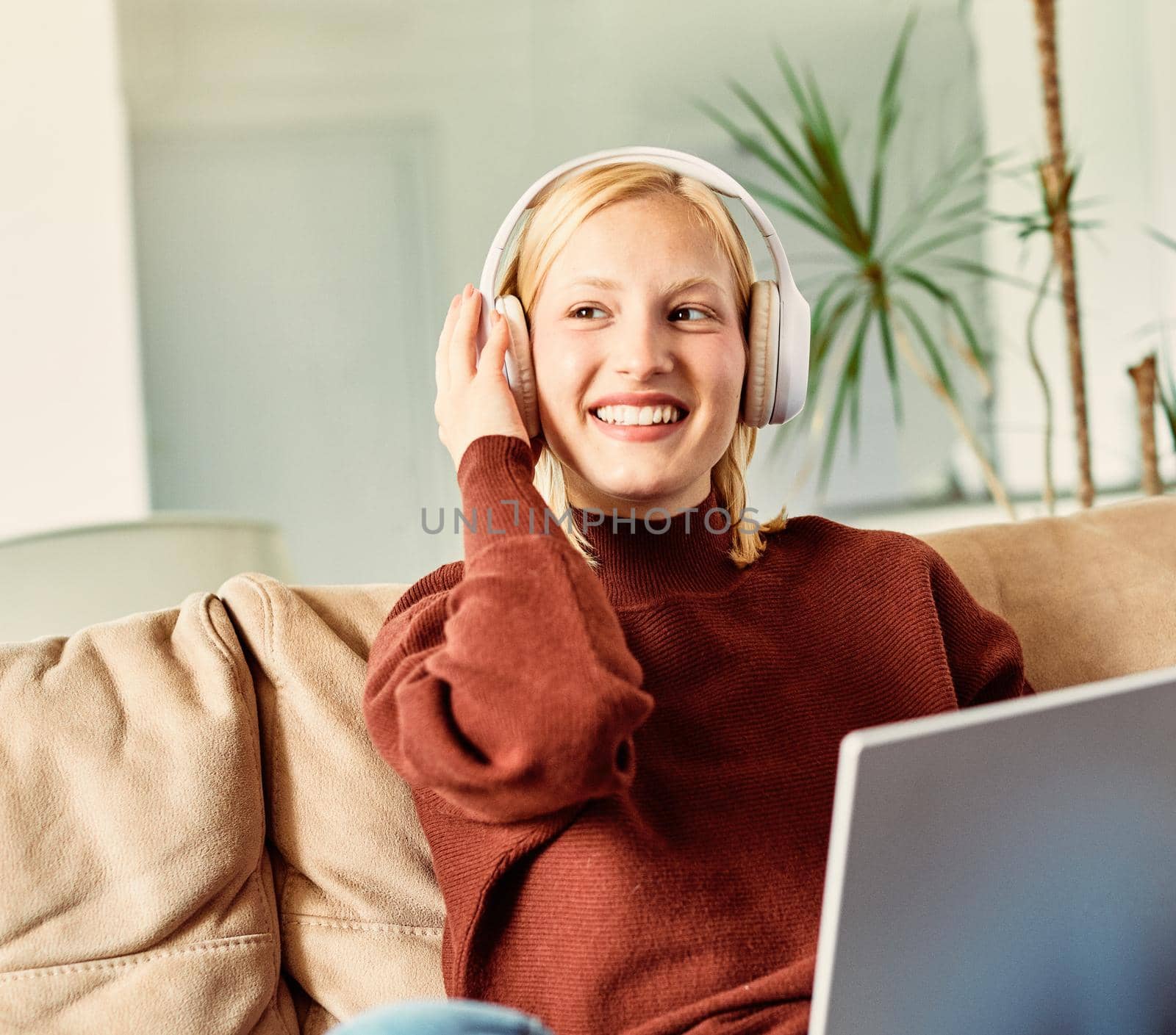 Portait of a young woman with headphones listening music on her laptop and relaxing on sofa at home