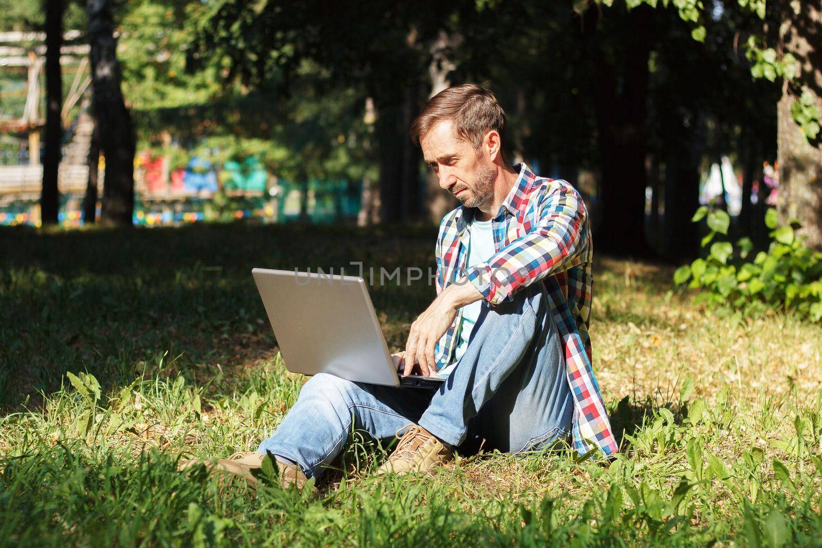 Adult man works on his computer in the park on the lawn.Remote work concept. The writer works remotely, enjoying nature.
