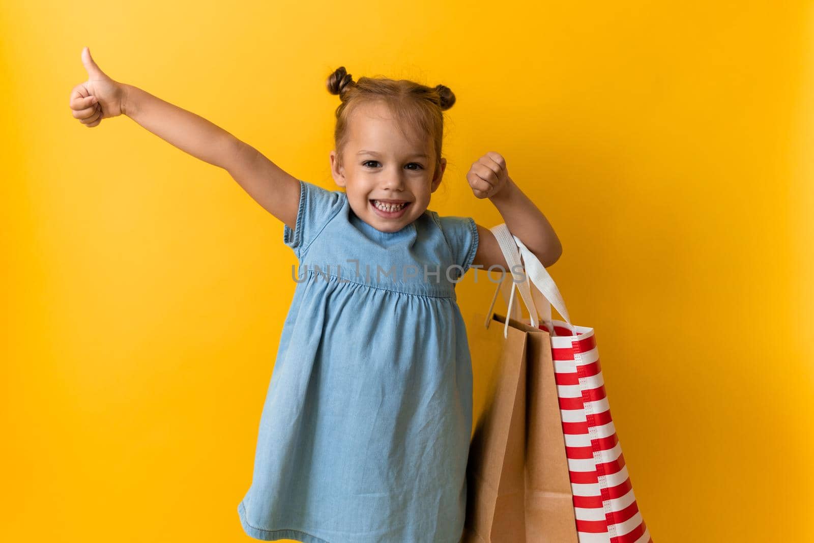 Portrait Caucasian Beautiful Happy Little Preschool Girl Smiling Cheerful And Holding Cardboard Bags Thumb Up Isolated On Orange Yellow Background. Happiness, Consumerism, Sale People shopping Concept.