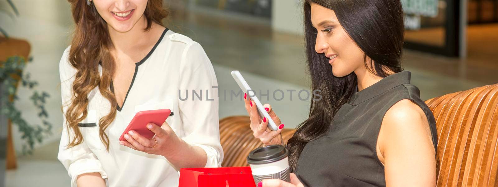 Two young caucasian women looking in smartphones smiling and sitting on the bench in a shopping mall