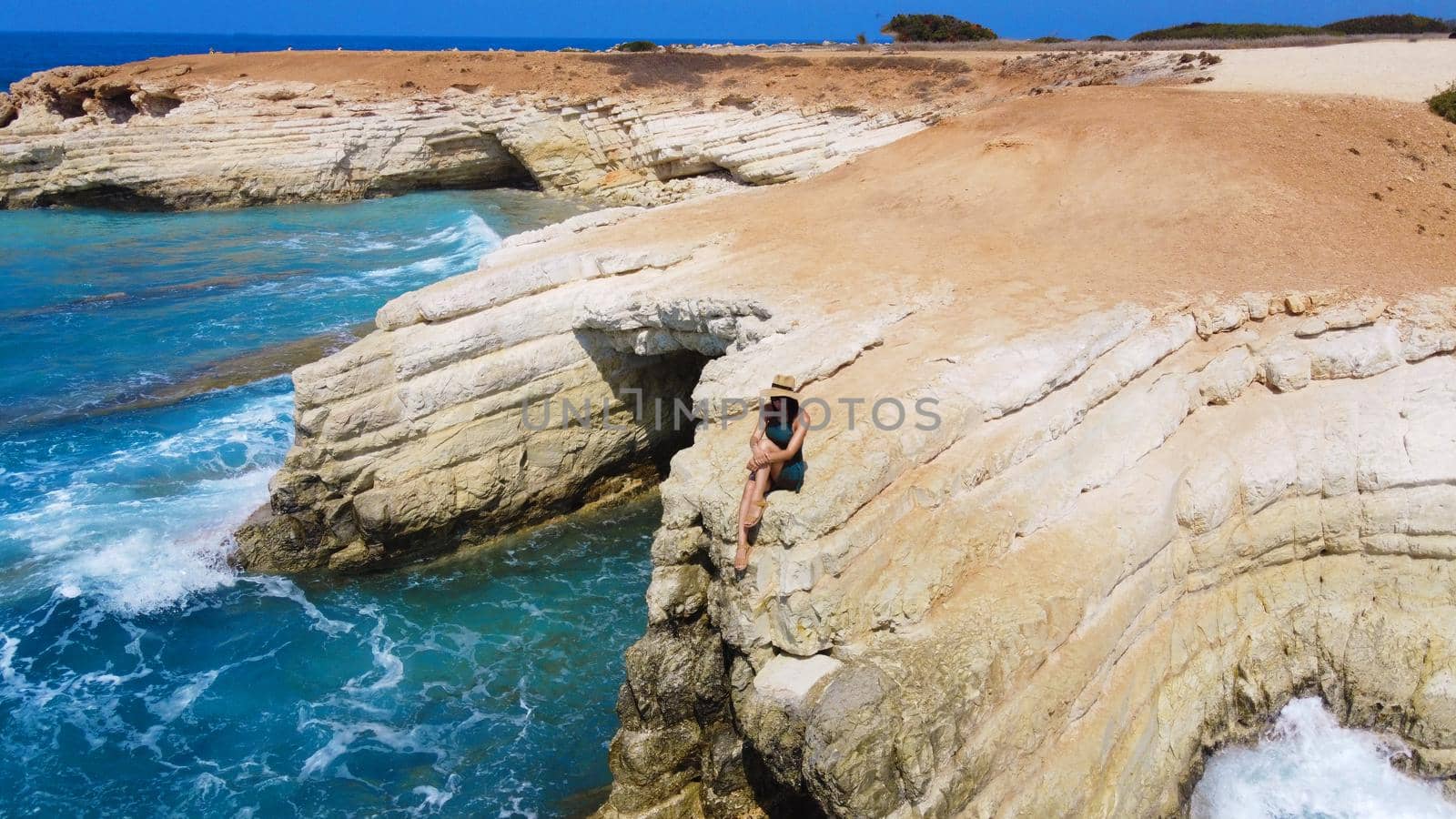 Beautiful girl sitting on a high rock and looking out to sea. top view. Girl in Hat on the edge of cliff. blue sea and high cliffs. Seascape. Girl at sunset. Sea tour. Blue sea, rocks