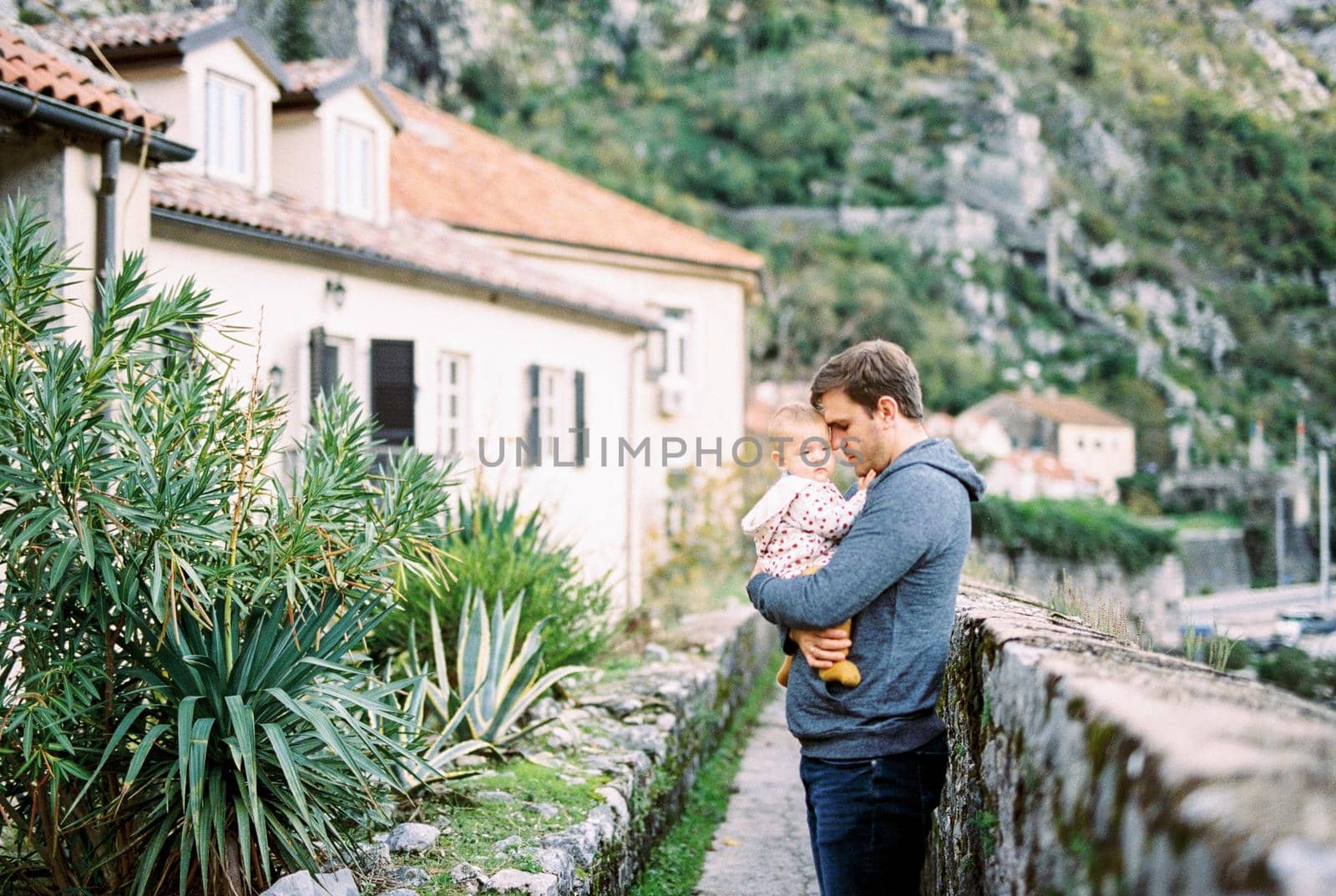 Dad with a baby in his arms stands near a stone fence in front of an old house at the foot of the mountains. High quality photo