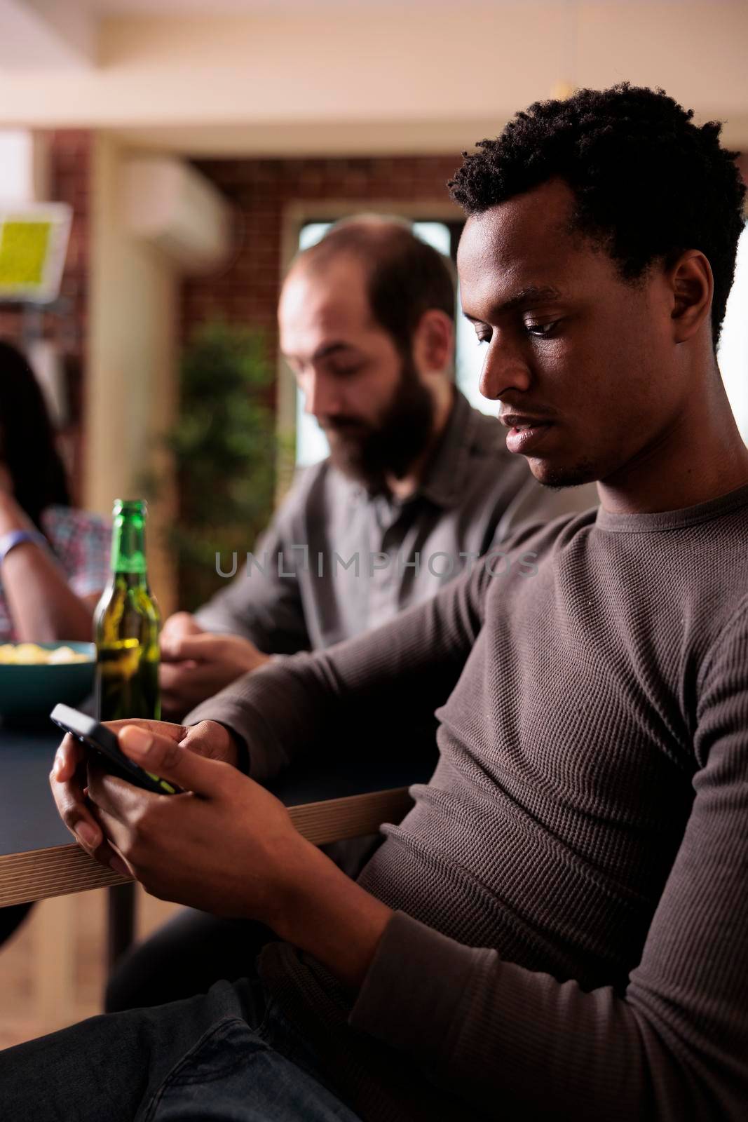 African american man with modern touchscreen smartphone sitting at table while browsing internet. by DCStudio