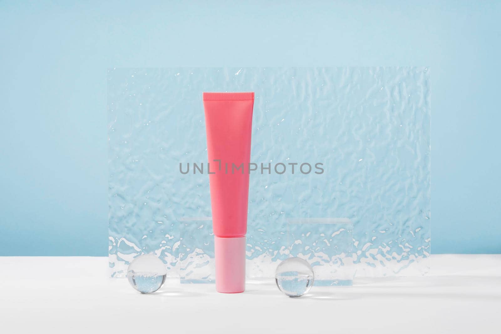 Unbranded cosmetic cream pink plastic tube mockup on blue background with stylish props, glass balls and acrylic plate. Lotion, concealer or beauty product packaging. Product presentation mock up